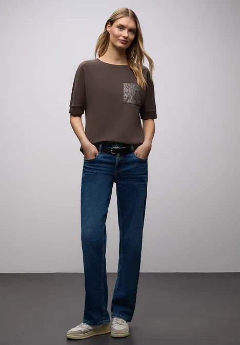 Eine Person trägt ein kurzes Taupe-T-Shirt mit Brusttaschenmuster, weite blaue Jeans und weiße Sneakers.